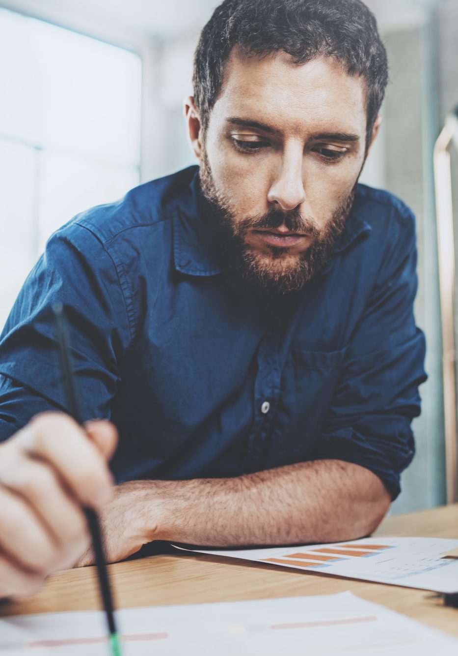 Young handsome businessman working contemporary notebook while sitting at the wooden table at sunny office.Man holding pencil hand.Blurred background. Horizontal.