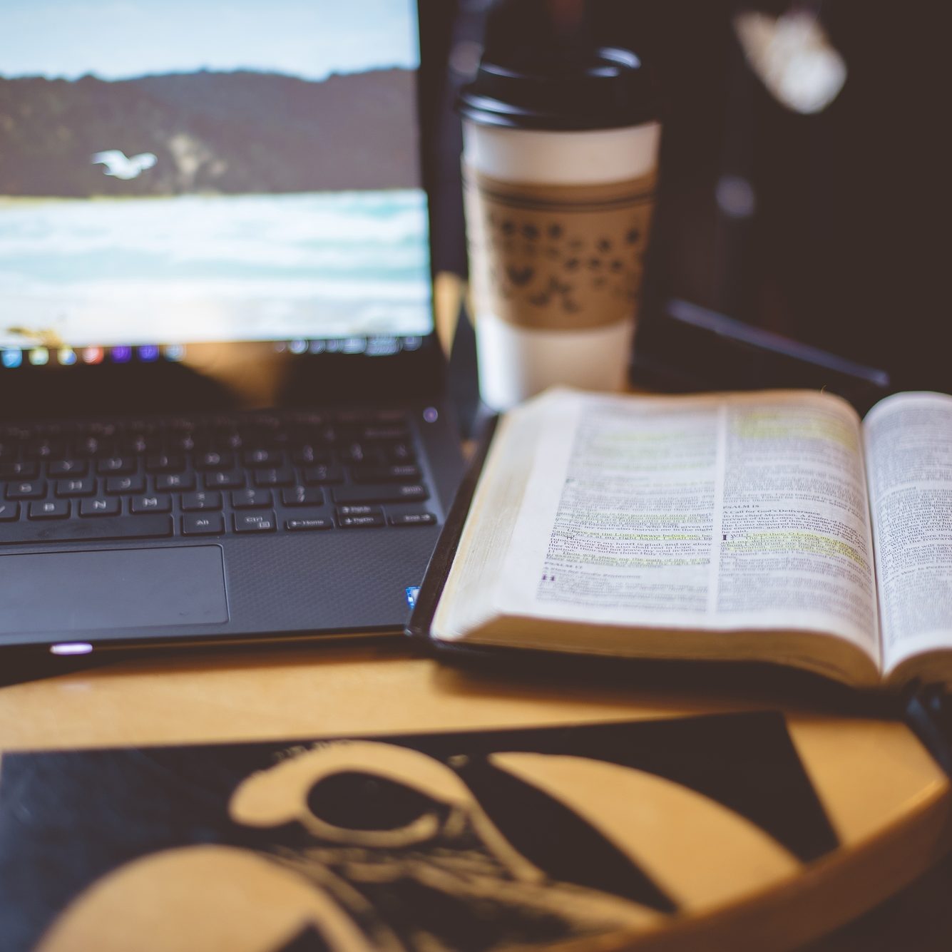 A selective focus shot of an open bible near a laptop and coffee on the table with a blurred background